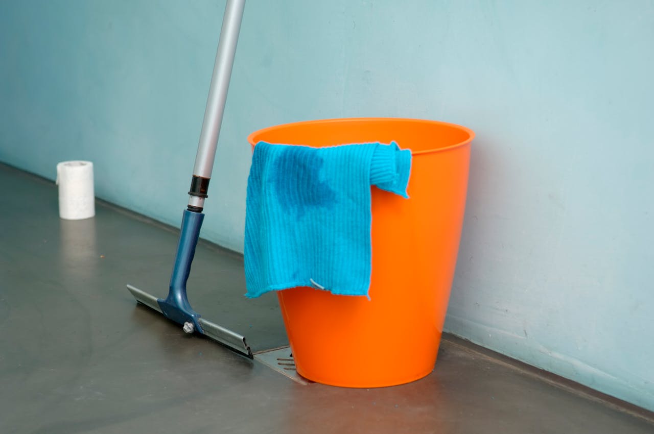 about-01 Close-up of cleaning supplies including orange bucket, mop, and blue rag indoors.